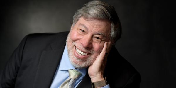 In a portrait with a dark studio background, Steve Wozniak leans forward, propping his face on his hand, smiling into the camera.
